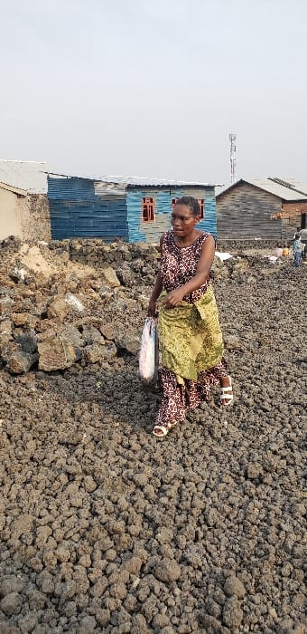 An adult female walks across uneven, dark grey, clumpy terrain in Buzove, Democratic Republic of the Congo. She is positioned slightly right of center, moving towards the viewer's right. She wears a sleeveless, patterned long dress in dark brown and light brown, with a green-yellow patterned fabric wrapped around her waist, and white sandals. In her left hand, she carries a white translucent bag containing a large, light-colored fish. To her immediate left is a mound of larger, darker rocky debris. In the background, multiple rudimentary structures are visible: a light-colored building with a corrugated roof on the far left, a central building constructed of blue corrugated metal with reddish-brown window frames, and to its right, a wooden plank structure with a corrugated roof. A tall communications tower stands in the far background to the right of the blue building. Two indistinct figures are visible further right in the distance near the background structures. The sky is uniformly overcast.