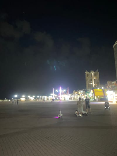 This is a night-time, wide-angle, low-resolution image of an outdoor promenade with cobblestone paving in the city of Merchanskoe, Russia. The sky is dark and cloudy, with some faint lights visible. In the foreground, there are two Segways, one upright and one on its side with a person lying on top of it. Two men are standing nearby, one wearing a light-colored shirt and shorts, the other in a dark shirt and shorts. Further back, there are several people walking and standing around various illuminated structures, including what appears to be a small amusement area with colorful lights. A tall, multi-story building with lights on is visible in the background, as well as other buildings with illuminated storefronts.
