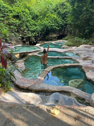A woman in a leopard print bikini stands in a series of tiered, natural-looking turquoise pools nestled within a lush, green jungle environment. She is facing away from the camera, with her right arm raised. The pools are constructed with rough, sandy-colored concrete borders and feature some decorative mosaic tiles on the bottom of some of them. Dense foliage, including large-leafed plants and trees, surrounds the pools, creating a secluded and tropical atmosphere. The scene appears to be a natural hot spring or thermal bath area in the city of Khlong Thom Nuea, Thailand. Sunlight filters through the trees, casting dappled shadows on the ground and water.