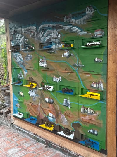 The image shows a beehive painted with a landscape depicting mountains, rivers, and various buildings such as churches and castles. The beehive is located outdoors, with visible brick pavement in the foreground and greenery in the background, suggesting a rural or garden setting in Dolenje Grčevje, Slovenia. The beehive has multiple individual compartments, each with a small entrance. Clusters of bees are visible in front of some of these entrances, particularly on the colored landing boards. The overall condition of the beehive appears to be well-maintained, with the painted artwork being a prominent feature.