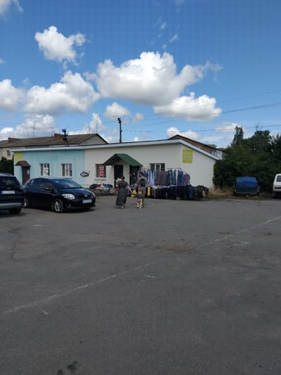 In Chudniv, Ukraine, on a bright, cloudy day, a street market is set up in front of a light blue and white building with a green awning. Clothes are displayed on racks outside the building, and a few people are browsing. A black Toyota Auris is parked in the foreground, and a black SUV is parked to its left. In the background, there are trees and more buildings. A blue tarp-covered trailer is visible to the right of the building, and a white van is parked further down the street.