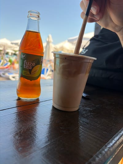 The image captures a close-up view of two drinks on a dark wooden table. To the left, a glass bottle of Fuze Tea, Lemon Lemongrass flavor, is visible, with its amber liquid and clear glass bottle catching the sunlight. The bottle's label features green and yellow accents. To the right, a clear plastic cup contains a layered beverage, likely iced coffee or a similar drink, with a brown frothy layer on top and a lighter layer below. A dark straw with a wooden textured section is inserted into the cup, and a hand with manicured fingernails is holding it. The background suggests an outdoor setting, possibly a beach or poolside, with glimpses of white umbrellas, lounge chairs, and a bright blue sky. The overall scene conveys a sense of relaxation and refreshment. The location is identified as Vetren Dol, Bulgaria, suggesting a possibly sunny day in a vacation or leisure spot.