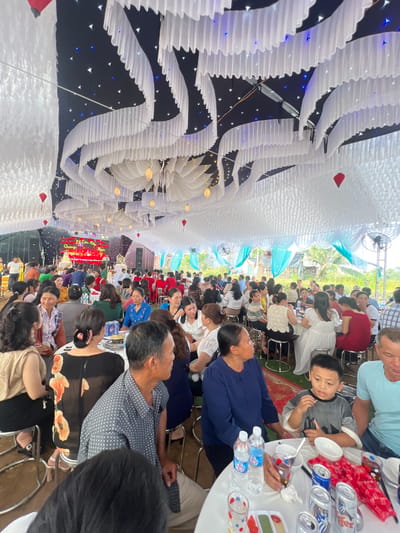 This media file captures a lively wedding reception held in a large, elaborately decorated tent or covered hall in Bình An, Vietnam. Numerous guests, comprising adults of various ages and some children, are seated at long tables, socializing and enjoying the festivities.

The most striking visual element is the overhead decoration, which features a dense arrangement of white, pleated fabric or paper structures, resembling clouds or stylized lanterns. These are illuminated by countless small, twinkling blue and white lights that mimic stars against a dark ceiling, creating a celestial and festive atmosphere. A few red, heart-shaped ornaments are also integrated into the decor. Further down the hall, large ceiling fans and some teal-colored translucent decorations are visible, suggesting an open-air or semi-outdoor setting.

Guests are seated at tables covered with white tablecloths. On the tables, bottled water, various brands of soft drink cans (such as Coca-Cola and 7-Up), and small plates with snacks or food are present. People are seen conversing, eating, and observing the event. A young boy in a grey shirt is visible near the foreground, looking towards the camera, while most attendees are oriented towards the far end of the hall.

At the distant end, a brightly lit stage or banner forms the focal point of the event. The banner, adorned with red and gold decorations, displays Vietnamese text. The legible text includes "Lễ Tân Hôn Ngọc" (Wedding Ceremony of Ngoc), "Quang," and "Ngày 8 tháng 3 năm 2023" (March 8, 2023), confirming the event as a wedding celebration for Ngoc and Quang on that date. The overall mood appears joyful and celebratory, with guests relaxed and engaged in the occasion. The lighting suggests the event might have transitioned from daytime into the evening.