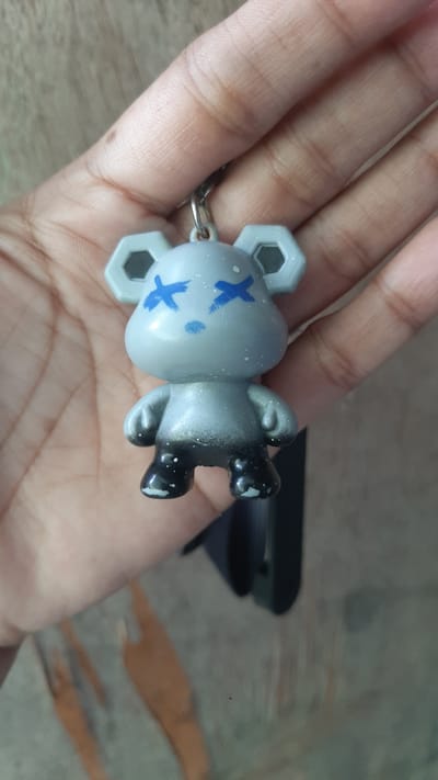 A close-up shot shows a hand holding a small, gray, cartoonish figurine with blue "X" marks for eyes and a small blue mark for a nose. The figurine has hexagonal ears, a rounded head, and a stout body with short arms and legs. Its lower legs and feet are painted black with white speckles. A metal keychain loop is attached to the top of its head. The background appears to be a textured wooden surface, possibly a table or wall, with some blurry shapes and lines suggesting it is indoors. The image is taken in Kawit, Philippines.