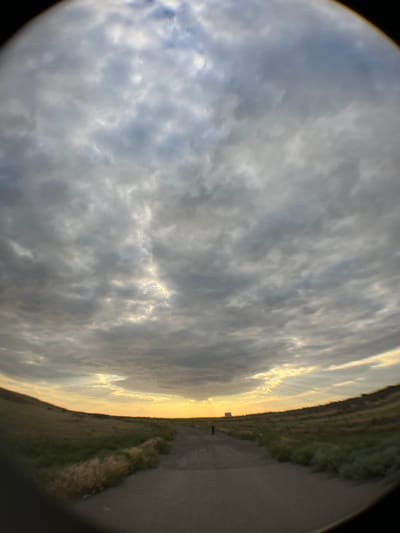 This fisheye lens image captures a dramatic sky over a rural landscape. The sky is filled with a mix of gray and white clouds, with bright rays of sunlight breaking through from the right side of the frame, suggesting a sunset or sunrise. The light creates a warm, golden glow along the horizon and illuminates the edges of the clouds.
To the right of the sky is a landscape that appears to be a dirt road or pathway running alongside a grassy embankment. The embankment is covered with dry grass and some green vegetation, suggesting a natural or undeveloped area. A single, dark silhouette of a person can be seen standing on the pathway in the middle distance, appearing small against the vastness of the landscape and sky.
The overall scene conveys a sense of solitude and the raw beauty of nature. The time of day is likely late afternoon or early evening due to the angle and color of the sunlight. The weather appears to be partly cloudy, with the sun intermittently piercing through the overcast. The location cues, such as the dirt road and the type of vegetation, are consistent with a rural setting in Russia, as indicated by the provided context. There is no visible text in the image.