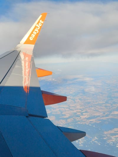 An aerial view captures the wing of an EasyJet airplane, its orange and white livery prominent against a backdrop of clouds and a patchwork landscape below. The wing, primarily dark blue and metallic gray, dominates the foreground. The EasyJet logo is clearly visible on the wingtip, written in white text on an orange background. The plane appears to be in flight, with the ground visible in the distance. The landscape below consists of agricultural fields, suggesting a rural or semi-rural area. The weather appears partly cloudy, with patches of blue sky visible between the clouds. There are no people or animals visible in the image, only the airplane wing and the landscape. The scene is static, depicting a moment in time during an aerial journey.