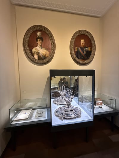 The image captures a scene inside what appears to be a museum or historic building, likely located in Sintra, Portugal. Two oval portraits of a man and woman in historical attire hang on the wall above three glass display cases, each showcasing different artifacts. The central display features silver boat models. The display on the left contains what seems to be illustrated manuscripts, and the one on the right has a wooden box. Reflections in the glass of the middle display reveal two people viewing the exhibits. The environment suggests a location with cultural or historical significance.