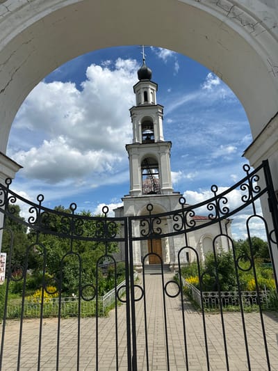 The image captures a view through a large, weathered arched gateway towards a white bell tower and a church in the distance. The gateway is made of white, cracked plaster. A black wrought-iron gate, decorated with ornate scrollwork and circles, is partially open in the foreground, obscuring a paved walkway leading to the church.
The bell tower is tall and slender, painted white, with multiple tiers and arched openings. It is topped with a dark dome and a cross. The church itself is also white, with classical architectural details, including columns and arched windows. A red-tiled roof is visible behind the bell tower.
The area in front of the church appears to be a garden with lush green trees and bushes. Patches of bright yellow flowers are visible amongst the foliage. The sky above is a vibrant blue, dotted with fluffy white clouds. The scene is bathed in bright daylight, suggesting a sunny day.
The location is identified as the Church of the Resurrection in the village of Yerynino, Russia. There are no people visible in the image.