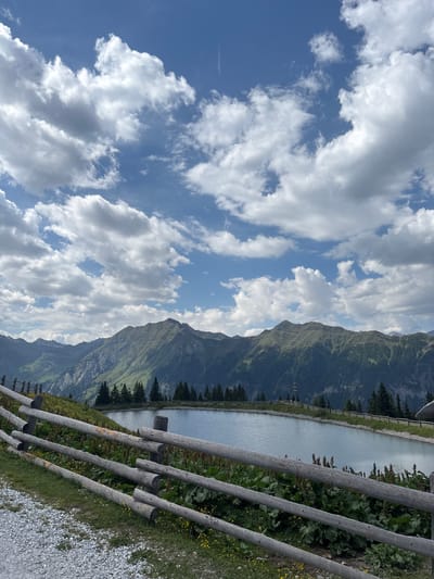 The image depicts a scenic mountain landscape in Ratschings - Racines, Italy. The scene is dominated by a vast expanse of sky filled with dramatic, fluffy white clouds against a clear blue backdrop. In the middle ground, a calm body of water, likely a reservoir or lake, stretches horizontally, bordered by lush green vegetation and a dark line of evergreen trees. The background is defined by a majestic mountain range, its slopes covered in green foliage and rocky outcrops, receding into the distance. A rustic wooden fence runs diagonally across the foreground, made of rough-hewn logs, separating the viewer from the natural scenery. The ground beside the fence is covered in gravel and green grass dotted with yellow wildflowers. The overall impression is one of serene natural beauty, suggesting a clear, pleasant day, possibly late morning or early afternoon given the bright sunlight illuminating the clouds and landscape. There are no people or specific activities visible, implying a peaceful, undisturbed natural setting. Some faint text is visible on the wooden fence posts, but it is illegible.