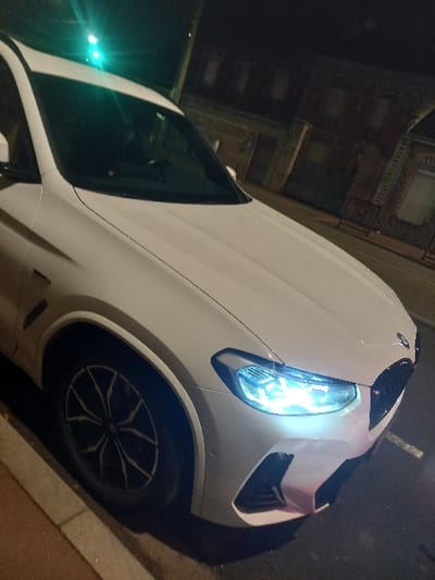 A white BMW X3 sits parked at an angle on a street with a sidewalk. The car's front is clearly visible, with its headlights shining brightly. The tires and rims are also prominent, and the background includes a building with windows and a street setting. The angle is a close-up shot.