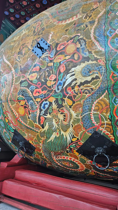 The image displays a close-up view of a large, ornately painted drum situated in Godang-ri, South Korea. The drum is adorned with vibrant, traditional Korean artwork featuring dragons, clouds, and floral motifs. The colors are rich and varied, including gold, red, blue, green, and black. Two black metal rings, each with a lion head design, are attached to the drum, presumably for handling or support. The drum rests on a weathered, red wooden stand. In the background, a portion of a decorated ceiling with patterned tiles is visible. The overall scene suggests a cultural or historical artifact displayed in an outdoor or semi-outdoor setting.
