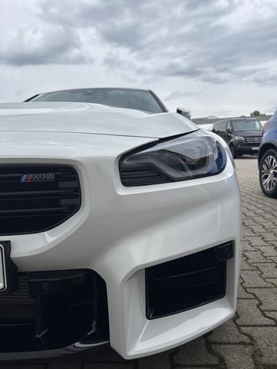 This media file shows a close-up, low-angle, vertical shot of the front of a white BMW M2 parked on a paved area with a cobblestone-like pattern. The car's headlight and grill are prominently featured. The grill has the "M2" badge with the iconic M colors. To the left of the car, a portion of a dark-colored van is visible, and behind it, the wheel of a blue vehicle can be seen. The sky is overcast with dramatic clouds.
The scene appears to be outdoors, possibly a dealership or parking lot, given the presence of multiple vehicles and the paved ground. The time of day is likely daytime, but the cloudy sky suggests it might be overcast or nearing evening. There are no people visible in the frame. The focus is entirely on the car's front fascia and the surrounding environment. The image is sharp and detailed, highlighting the car's design elements and the texture of the ground. No specific activity is depicted, suggesting the car is stationary.
The location context provided is Wettstetten, Germany.