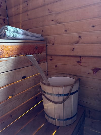 In an interior scene, a wooden bucket filled with water and a ladle sits on a wooden bench within a sauna. The walls and ceiling of the sauna are constructed from horizontal wooden planks. A neatly folded light-colored towel is placed on a higher wooden shelf. The lighting in the sauna is warm and inviting, with visible yellow light emanating from between the wooden slats. The overall environment suggests a relaxing and traditional sauna experience. The location of this scene is in the city of Семилуки, Russia.