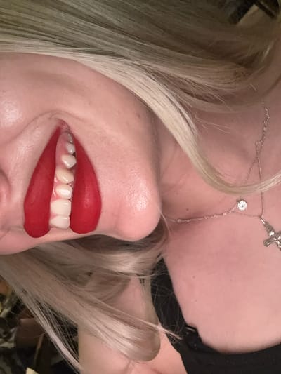 The image displays a close-up, angled view of an unidentified individual's head and upper torso, oriented approximately 90 degrees clockwise from an upright position. Long, light-colored hair, predominantly blonde, cascades across the upper and lower portions of the frame. The individual's skin tone appears fair. A prominent feature is the individual's mouth, adorned with bright red lipstick. The lips are visibly stretched horizontally, exposing the upper dental arch, including several white, aligned teeth and portions of the upper gums. The lipstick forms distinct, elongated red shapes on either side of the exposed dental area. Around the neck/collarbone area, a silver-colored necklace is visible, featuring two distinct pendants: a small, round, clear faceted stone and a detailed crucifix. A segment of black fabric, consistent with clothing, is visible along the lower right edge of the frame. The background is dark and out of focus. The lighting suggests an indoor setting. This scene is situated in the city of Strumica, North Macedonia.
