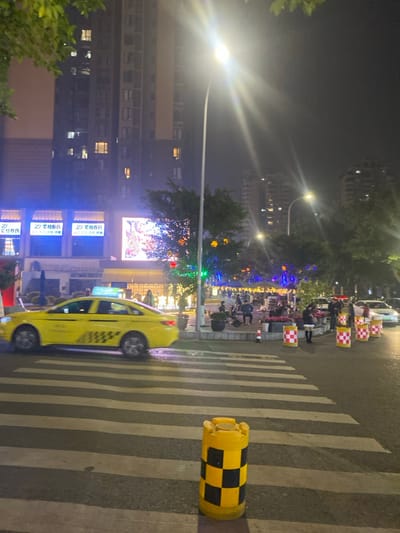 This nighttime image depicts an urban street scene in 天宫殿街道, China. A yellow sedan taxi, identifiable by a light blue roof sign and black checkered decals on its rear doors, is in motion, driving from left to right across a white-striped pedestrian crosswalk. In the immediate foreground, centered on the lower right, a yellow and black checkered cylindrical bollard stands on the asphalt.
To the left, a multi-story building with illuminated windows rises into the dark sky. Below it, at street level, are commercial establishments featuring bright blue illuminated signs, two of which display the characters "荣柏春氏," adjacent to a large, brightly lit digital billboard showcasing vibrant, abstract patterns.
A wide sidewalk and public gathering area are visible to the right of the street. This area is adorned with trees wrapped in colorful string lights (blue, red, orange, green) and features multiple red and white checkered barriers or planters along its perimeter. Numerous individuals are present in this public space, walking, standing, and congregating around various points, some illuminated by overhead lights suggesting stalls or seating areas. One person, wearing a dark jacket over a light-colored top, is visible near the checkered barriers on the right. Several parked vehicles are discernible further down the street on the far right. The scene is brightly lit by multiple streetlights, which produce prominent light flares, and by various commercial and decorative illuminations.