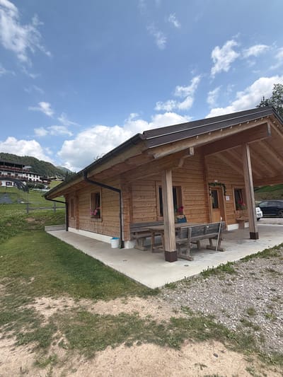 The image shows a rustic wooden cabin situated in a mountainous, rural area. The cabin has a covered porch with wooden tables and benches. Two windows on the front of the cabin are adorned with flower boxes. A white bucket and a broom are visible near the entrance. The surrounding environment includes a grassy lawn, a gravel path, and a hillside with trees and buildings in the background. The sky is blue with scattered white clouds. The scene appears to be in Lateis / Latais, Italy, as indicated by the architecture and landscape.