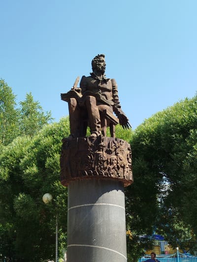 The image shows a bronze statue of a seated man reading a book, positioned on top of a cylindrical pedestal. The pedestal is decorated with bas-relief carvings of figures and scenes. The statue is located in the city of Yalutorovsk, Russia, in a public area with lush green trees and a clear blue sky. The lighting suggests it is daytime, with the sun casting shadows. In the background, a street lamp is visible amongst the foliage. Further in the background, some playground equipment and a blue fence can be seen, and a person is partially visible near the bottom right corner.