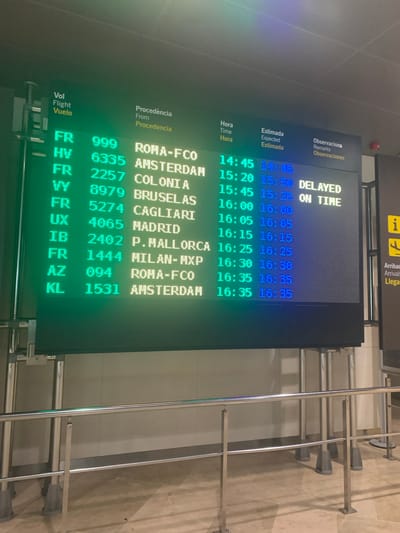 The image shows a digital flight information board at an airport. The board displays flights with their origin, flight number, departure time, estimated departure time, and remarks. The board is illuminated with green and white text. To the right of the flight board, there is a sign with an "i" symbol and an airplane symbol. The environment appears to be an indoor public space, possibly a terminal. The perspective is from a low angle, looking up at the board. The lighting is bright, suggesting artificial illumination. The floor is tiled. There are metallic stanchions and railings in the foreground. The image was taken in Manises, Spain, likely at the Valencia Airport (VLC), as indicated by the flight information often seen at this airport.