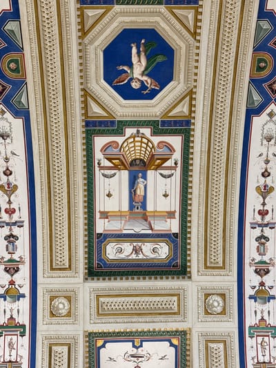 The image displays a highly detailed, ornate painted ceiling, likely an architectural fresco, found in San Lorenzo de El Escorial, Spain. The ceiling is divided into numerous geometric panels and borders, featuring a rich color palette of blues, golds, whites, reds, and greens.

Dominating the upper central octagonal panel is a fresco of a winged cherub, depicted in an upside-down pose, with green drapery trailing behind it. Other panels showcase intricate decorative motifs, including geometric patterns, stylized floral elements, and trompe-l'œil architectural details. Several rectangular panels contain depictions of mythological or allegorical figures, often rendered in a classical style, standing within architectural niches or elaborate frames. These figures include a woman in a yellow garment and a male figure in a warrior-like stance. Smaller details include representations of birds, vases, and what appear to be decorative lamps or urns. The overall impression is one of opulence and refined artistic execution, consistent with Renaissance or Baroque decorative schemes found in palatial or religious structures.