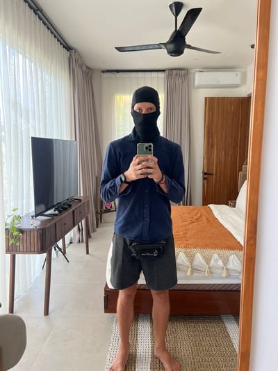 A man wearing a black balaclava takes a selfie in a mirror. He wears a dark blue button-down shirt and grey shorts. He holds a phone with a green case in his right hand. A black bag hangs around his waist. The reflection shows a bed with a brown and white patterned blanket, a wooden door, a black ceiling fan, and an air conditioning unit. The foreground contains a television on a wooden stand, with a plant on the stand. Behind the television, white blinds cover a window. The floor is light-colored tile. The man is standing indoors in what appears to be a residential setting, possibly in Buduk, Indonesia.