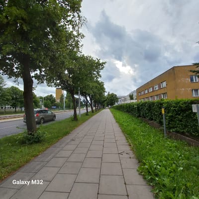 This is a full-frame, eye-level, outdoor, wide-angle photograph taken on a cloudy day in Olsztyn, Poland, likely during daytime. The scene depicts a paved pedestrian walkway lined with trees on the left and a dense green hedge with a yellow building behind it on the right. A street with tram tracks and a car is visible to the left of the walkway. The walkway is made of grey square paving stones, some of which show wear and tear. The grass verge to the right of the walkway is overgrown with various plants. The sky is filled with heavy grey clouds, with some lighter patches suggesting diffused sunlight. The yellow building on the right appears to be an apartment or office building with multiple windows. The overall atmosphere is that of a residential or urban street with public amenities. The bottom left corner of the image displays a watermark that reads "Galaxy M32".
