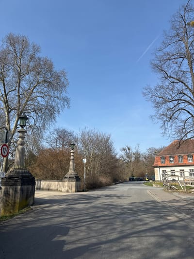 The image depicts an asphalt road leading into the Kurpark area of Bad Nauheim, Germany, on a clear, sunny day. The scene is devoid of people or active vehicles.
In the foreground, the asphalt road displays multiple long, dark shadows cast by unseen elements to the left, indicating a low sun angle. Two circular manhole covers are embedded in the road surface.
The mid-ground features a bridge entrance marked by two ornate, fluted stone pillars, each topped with a dark, multi-paned lantern-style lamp. These pillars flank the road, connected by a stone bridge wall. To the left of the left pillar, a red-bordered circular sign with "12t" in black indicates a weight limit, and below it, a white rectangular sign displays "E13." To the right of the right pillar, a light blue rectangular sign with a white arrow is visible.
The environment predominantly features deciduous trees with bare branches, suggesting late winter or early spring conditions. Dense brown scrub and smaller trees line the sides of the road beyond the bridge structures. A large clump of mistletoe is visible in a tree on the right side of the frame.
On the far right, a two-story building with a light-colored facade, numerous white-framed windows, and a red-tiled hip roof is visible. A rustic wooden fence runs in front of this building. Further down the road on the right, a yellow sign with black text is partially discernible.
The road continues into the background, where several dark-colored trash bins are situated on the left. The sky is a uniform, clear blue, traversed by one prominent white contrail from an aircraft.