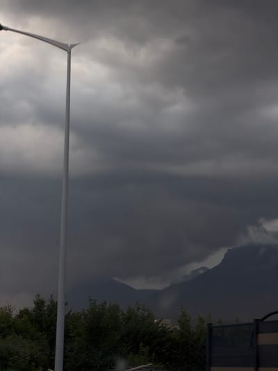 The image depicts a landscape scene under a dark, cloudy sky. A tall, white street lamp stands prominently on the left side of the frame, its arm extending horizontally to the left. In the background, dark, silhouetted mountains are visible, partly obscured by heavy clouds. Below the mountains, a line of dark green trees stretches across the lower portion of the image. The overall atmosphere is somber, suggesting overcast or stormy weather. The location is identified as Groisy, France.