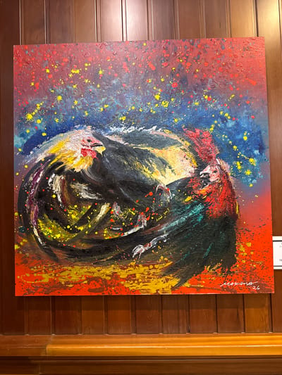This is a photograph of an abstract painting featuring two roosters in a circular motion against a vibrant, speckled background of red, blue, and yellow. The painting is hung on a dark wooden paneled wall. The roosters are depicted with energetic brushstrokes, with dominant black and red colors, and splashes of yellow and white on their heads and tails. The background is textured with a splatter effect of yellow and red dots on a gradient of blue to red. The signature "MAKM 26" is visible in white on the lower right corner of the painting. A small white card is partially visible on the right edge of the frame, suggesting the painting is on display in a gallery or exhibition space, potentially in Torrejón de Ardoz, Spain. The painting is illuminated by artificial light, casting subtle shadows.