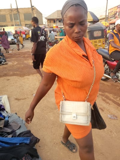 A woman in an orange top with a white purse stands in an outdoor market area. She has a headscarf and wears sandals. Nearby is a pile of clothing on the ground. Behind her, blurred figures and vehicles suggest a bustling scene. Buildings are visible in the background, indicating an urban environment. The ground appears to be dusty.