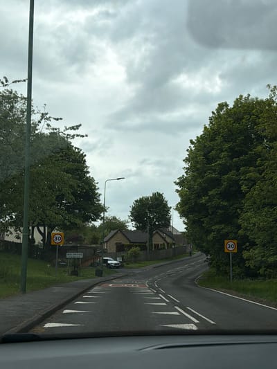 A street scene in Waldridge, a village in the city of Durham, United Kingdom, is captured from the perspective of a vehicle. The overcast sky suggests it could be a typical day in the region. The road is marked with white dashed lines towards the center and solid white lines with adjacent chevrons along the curb, indicating lane markings. Two prominent yellow circular road signs reading "30" signify a 30 miles per hour speed limit. Lush green trees line the roadside. Buildings, including houses, are partially visible behind the vegetation. A street sign reading "WALDRIDGE" is also visible, confirming the location. A silver car is parked along the road. A lamp post stands along the road.