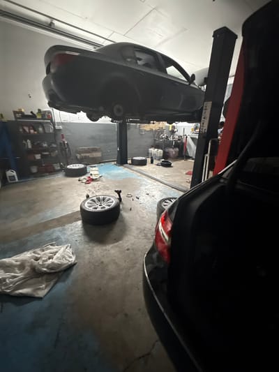 In this image, a dark-colored car is elevated on a hydraulic lift in what appears to be a dimly lit auto repair shop. The car is positioned with its underside facing the camera, revealing its suspension components. Multiple car tires, some mounted on rims with silver spokes, are scattered on the concrete floor. One tire is prominently featured in the foreground, angled towards the viewer. In the background, shelves are filled with various containers and tools, suggesting a workspace for mechanics. A white cloth or rag is draped on the floor to the right of the frame. The overall scene depicts a car undergoing maintenance or repair. The lighting suggests it is likely nighttime or indoors, and there are no visible weather cues. The location is identified as Čelákovice, Czechia.