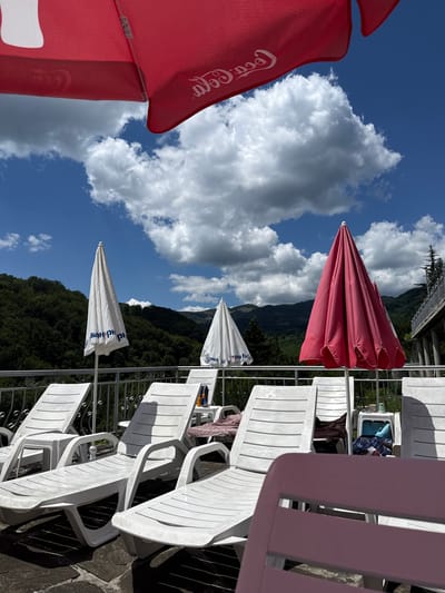 In Shipkovo, Bulgaria, an outdoor scene unfolds under a bright blue sky dotted with fluffy white clouds. In the foreground, several white plastic lounge chairs are neatly arranged on a stone patio. Some of these chairs are occupied by dark blankets and towels. A light pink plastic chair is positioned in the very front, partially obscuring the view of the other chairs.
Behind the lounge chairs, a white metal railing runs horizontally, separating the patio area from a lush, tree-covered hillside that rises in the background. Several closed umbrellas are visible: two white ones with blue lettering, and one bright pink umbrella to the right. Above these, a large red umbrella, branded with the Coca-Cola logo, is partially visible, suggesting it is a prominent feature of this outdoor space. The overall impression is one of a peaceful, sunny day at a resort or a similar recreational area.