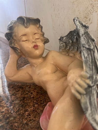 This media file contains a close-up, sideways view of a statue resembling a cherub or angel. The statue is rendered in a realistic style, with painted details and a light skin tone. It appears to be made of a material like resin or plaster.
The subject is positioned on its side, with its head tilted back and resting on its arm. The eyes are closed, and the mouth is slightly open with painted red lips. The hair is depicted with dark, wavy strands. There are hints of wings or some sort of textured, rocky element behind the head and upper body of the statue, rendered in shades of gray and white. A faint pink element, possibly fabric or part of the statue's design, is visible near the lower right of the frame.
The statue is resting on a surface that appears to be a speckled, granite-like material. In the background, there is a light-colored, textured surface, possibly a wall or tile. The lighting suggests an indoor setting, and no specific time of day or weather conditions can be determined. There is no visible text within the image. The overall emotion conveyed by the statue's pose and painted features is one of rest or peaceful slumber.