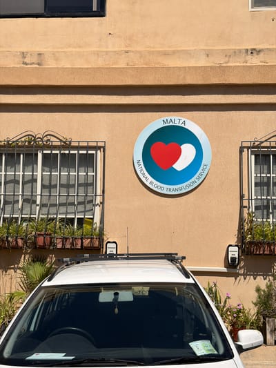 The image shows the exterior of the National Blood Transfusion Service building in Pietà, Malta. The building has a beige-colored facade with horizontal architectural elements. Two windows with black metal bars and potted plants in front of them are visible. Above the right window, a circular emblem with a blue and white background is mounted on the wall. The emblem features the word "MALTA" at the top and "NATIONAL BLOOD TRANSFUSION SERVICE" in a curve below. Two hearts, one red and one white, are depicted within the emblem. In the foreground, the roof and windshield of a white car are visible, with a parking permit visible on the dashboard. The scene appears to be outdoors during the day, with shadows cast by the building and vegetation.