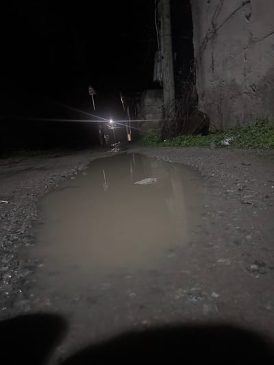 The scene depicts a dark, outdoor environment at night in Lehvaz, Armenia. The foreground is dominated by a large, irregularly shaped puddle of muddy water on an unpaved or heavily worn dirt/gravel path. The ground surrounding the puddle is uneven and composed of dark, granular material. To the right, a textured concrete or stucco wall with visible cracks and discoloration runs parallel to the path. At the base of this wall, green vegetation or weeds are present. A wooden utility pole is positioned adjacent to the wall, extending vertically out of frame, with associated wiring visible. In the mid-ground, a single, intense artificial light source, likely a streetlamp, emits a strong glare, creating a clear reflection in the puddle. This light partially illuminates a narrow passage or road extending into the darkened distance. To the left of the light source, a pole-like structure with horizontal appendages is faintly visible. The overall environment appears deserted, with no individuals or direct actions observed.