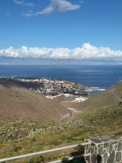 This image captures an elevated panoramic view of San Sebastián de la Gomera, Spain, nestled within a valley that opens to the Atlantic Ocean.
In the foreground, a gray metal guardrail with two horizontal bars is supported by a low stone wall composed of dark, irregularly shaped flagstones. Immediately beyond the railing, the terrain slopes downwards, covered in arid vegetation, including sparse green scrub and scattered succulent-like plants.
The midground features steep, rugged hillsides on both the left and right, predominantly displaying dry, reddish-brown earth with patches of green and grayish-green vegetation. A paved, dark asphalt road with white markings curves downwards into the valley from the lower center of the frame, leading towards the town. Another section of a winding road ascends the hillside on the far right.
The town of San Sebastián de la Gomera is situated in the central midground, a cluster of numerous low-rise buildings with varied roof colors such as red, white, and brown, and facades ranging from white to light earth tones. The town extends along the coastline into a bay, where a marina or harbor is clearly visible on the right, populated with multiple moored boats. A thin, prominent communication tower stands on the upper right hillside.
The background consists of the vast, deep blue Atlantic Ocean, extending to the horizon. Across the ocean, a faint outline of a distant landmass, consistent with the island of Tenerife, is visible. This distant land is positioned beneath a long, horizontal band of dense white cumulus clouds that stretches across the entire width of the sky. Above this cloud band, the sky is a clear, bright blue with a few scattered, wispy white clouds. The scene is brightly lit by natural sunlight, casting shadows that highlight the contours of the landscape. No people or active interactions are discernible.