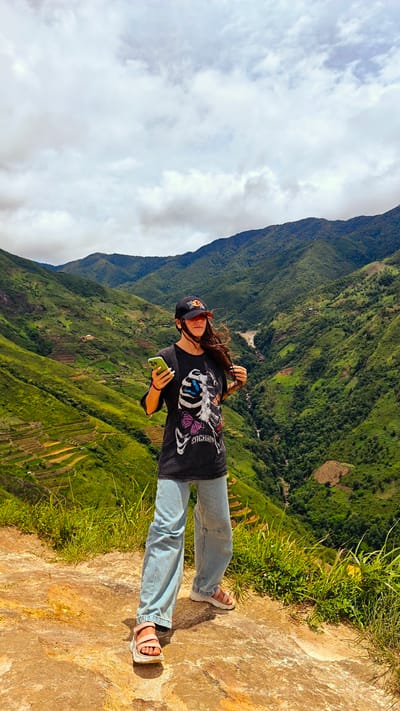 A woman stands on a cliff edge in Bắc Yên, Vietnam, holding a smartphone and looking towards the lush, terraced green mountains. She wears a black t-shirt with a skeletal design, light blue wide-leg jeans, a black baseball cap, and tan sandals. The background features dramatic mountain slopes with visible rice terraces, a winding river, and a cloudy sky. The terrain beneath her is rocky and earthen, with green grass growing at the edge.
