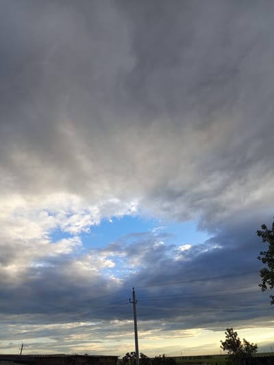 The media file shows a wide shot of a cloudy sky over a rural landscape. The sky is filled with a mixture of dark, heavy clouds and lighter, wispy ones, with patches of bright blue sky visible in the center. The lighting suggests it is either early morning or late afternoon, with a soft glow near the horizon.
In the foreground, there are hints of structures, including what appears to be a wooden building with a corrugated roof on the left. Several utility poles with wires stretch across the frame, indicating a presence of infrastructure. Trees are scattered across the landscape, with one prominent tree on the right side of the image, its leaves dark against the sky. The overall setting appears to be a rural or semi-rural area in Älimbet, Kazakhstan, characterized by open fields and minimal development.
The weather appears to be changeable, with the presence of both dark and light clouds, possibly indicating an approaching storm or the aftermath of one. There is no discernable activity or event taking place, and no visible text in the image. The mood is somewhat dramatic due to the heavy clouds, but the breaks of blue sky offer a sense of hope or calm.