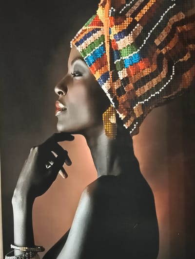 A close-up, profile view of a dark-skinned woman wearing a brightly colored, sequined headwrap and earrings. She is looking to the left with her chin resting on her hand. The background is dark and out of focus. There are no indications of location in the city of Константиновка, Russia.