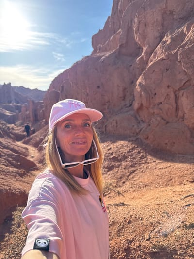 A woman poses for a smiling selfie outdoors, set against a backdrop of majestic reddish-brown rock formations and a bright blue sky. The image is taken from a slightly upward angle, with the sun visible in the lower left corner, indicating a clear, sunny day. The woman has long blonde hair and is wearing a light pink long-sleeved t-shirt and a matching pink baseball cap. White sunglasses are perched on her head, holding her hair back. A logo, possibly stylized letters like "NY," is visible on her cap. On the left sleeve of her t-shirt, a small black patch with a white circular logo, resembling a stylized peace sign, is visible. Her expression is happy and direct to the camera. The rugged, textured rock walls with varying shades of red and brown suggest a canyon or mountainous region, consistent with the provided location context of Tosor, Kyrgyzstan. Some wispy clouds are present in the sky.