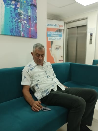 A man is sitting on a teal sofa in what appears to be a waiting room. He is wearing a white short-sleeved shirt with a blue dolphin pattern and dark green pants. His head is bowed, and he is looking down at his phone, which is lying on the sofa next to him. In the background, there is an elevator door and a poster with an image of an eye and text in Turkish. The walls are white, and there is abstract art on the left wall. The lighting is bright, suggesting an indoor setting. The overall impression is one of a person waiting, possibly at a medical clinic given the poster.
