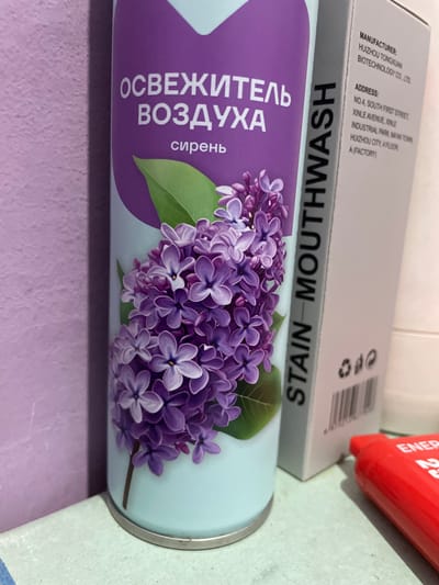 The image shows a close-up of a lilac-scented air freshener can next to a box of mouthwash. The air freshener can has text in Russian that translates to "AIR FRESHENER LILAC". The mouthwash box has text in English indicating it is "STAIN MOUTH WASH" and lists the manufacturer as "HUIZHOU TONGXUAN BIOTECHNOLOGY CO., LTD" and its address in China. A red object, possibly a lipstick or a small cosmetic item, is visible on the right side of the image. The scene appears to be indoors on a surface with a light green and blue textured finish. There are no people or animals present, and the environment is neutral and appears to be a retail setting or a shelf in a home. The city of Buynaksk, Russia, is not depicted in this image, nor is there any information in the image that suggests a connection to this location.