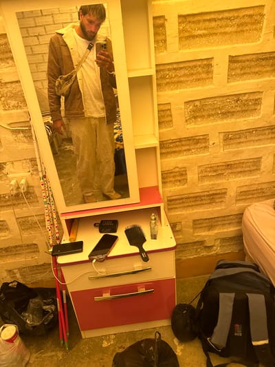 The image displays an indoor scene within a room in a salt hotel or guesthouse, indicated by the walls constructed from distinctly textured, light-colored salt bricks, common in Uyuni, Bolivia. A male individual, with light hair and a beard, is captured in a full-length mirror, taking a selfie with a smartphone held in his right hand. He is dressed in a brown, sherpa-lined jacket over a white t-shirt, light-colored textured trousers, and light shoes, with a beige crossbody bag worn across his chest. The mirror is integrated into a white and pink dresser that features two lower drawers and open shelving to the right of the mirror. On the dresser's top surface, from left to right, are a black rectangular object, a smartphone connected by a white charging cable, a black JBL portable speaker, a black hairbrush, and a clear glass perfume bottle. To the left of the dresser, on the floor, are black plastic bags, a white container, and several long, thin pink objects. To the right of the dresser, a blue and grey backpack with "ACS" branding is visible on the floor, positioned near a bed with a pink mattress. The floor is light-colored and textured.