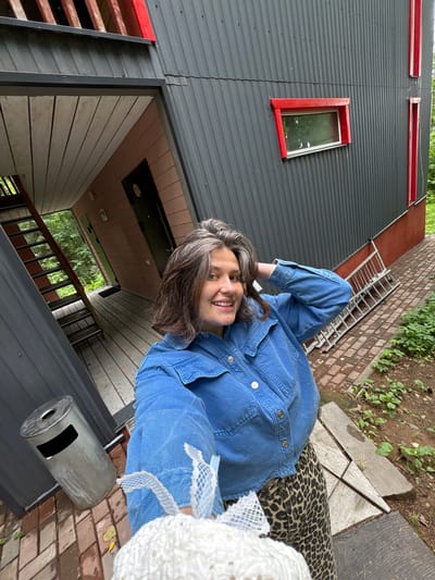 A woman with graying brown hair and a denim jacket smiles at the camera, holding a white object wrapped in netting. She is standing on a brick pathway next to a modern, angular building with dark gray corrugated metal siding and red accents around windows and doors. To her left, an external metal staircase leads up to a balcony. A silver trash can is visible near the staircase. In the background, green trees suggest a natural setting. The scene appears to be outdoors.