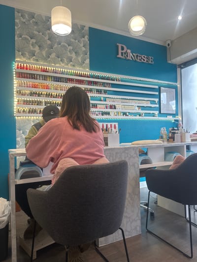 The image depicts the interior of a nail salon located in Malakoff, France. Two individuals are present, seen primarily from their backs. A customer, wearing a pink top and light pants, is seated in a dark grey upholstered chair with a pink cushion at their lower back, facing a white marble-patterned manicure table. Their dark hair is visible down their back. Opposite the customer, a nail technician in a dark baseball cap with a white "C" logo and an olive-green vest is partially visible, facing the customer across the table. A "SUNUV" brand UV nail lamp is positioned on the table between them.
The salon's back wall features a turquoise-painted upper section and a lower section covered with white wallpaper displaying a light blue ginkgo leaf pattern. Multiple white, illuminated shelves line the wall, meticulously displaying hundreds of bottles of nail polish. Above the nail polish display on the right, the word "PRINCESSE" is affixed to the wall in pink, stylized letters with a crown motif. A framed document hangs on the right wall. The ceiling is white with two cylindrical hanging light fixtures and a recessed light. A white security camera is mounted high on the right wall near a window or door frame. The floor is dark wood or laminate. On the right side of the image, another dark grey upholstered chair with a pink cushion is visible next to a second white marble-patterned table holding various nail care products, bottles, and tools, including additional UV lamps on a lower shelf. A black trash can is partially visible on the far left.