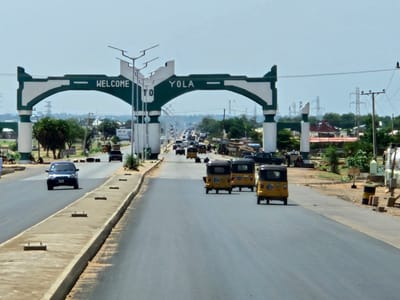 The image shows a wide asphalt road leading into the city of Yola, Nigeria, identified by a prominent green and white "WELCOME TO YOLA" archway spanning the road. A concrete median divides the road.  Several yellow auto rickshaws (tuk-tuks) are traveling towards the viewer. Further in the distance, other vehicles, including cars and trucks, are visible on the road. On the right side of the road, there are objects such as barrels, blocks, and posts. Along the sides of the road are trees, bushes, buildings, and power lines.  In the background, electrical towers are visible.