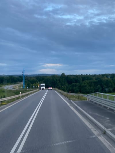 The image shows a road scene from the perspective of a vehicle driving on a highway. The road is paved with asphalt and has double white lines marking the lanes. There are guardrails on the sides of the road. In the distance, a white truck and a white car are visible, both driving in the same direction as the camera. The landscape on either side of the road consists of green trees and vegetation. The sky is overcast with dark blue clouds. A distinctive blue utility pole with multiple arms is visible to the left of the road, along with a curved exit ramp. The location is identified as Podłęże, Poland.