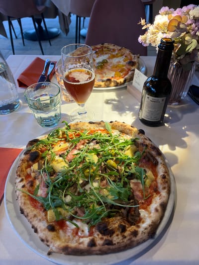 An overhead shot captures a dining table set for a meal at what appears to be an Italian restaurant in Sogliano Cavour. The scene is dominated by two pizzas, drinks, and decorative elements.

In the foreground, a large, freshly baked pizza with a visibly charred, puffy crust is presented on a white plate. It's generously topped with arugula, sliced ham, mushrooms, and artichoke hearts, along with melted cheese and tomato sauce. Partially visible in the background is another pizza with a different topping, possibly featuring yellow ingredients.

To the left of the main pizza, a stemmed glass holds an amber-colored beverage, likely beer, with some foam at the top. A clear glass, mostly filled with water, is placed next to it. Further back, a dark wine bottle, labeled "Calidore PRIMITIVA," stands upright, its label clearly showing "alc. 8,5% vol", "Product of Italy", and "2007/06-1525". A small wooden block bearing the word "Tavolo" (Italian for 'table') is positioned near the wine bottle.

The table is covered with a white tablecloth, and a folded orange napkin with cutlery underneath is visible. In the upper left corner, a decorative arrangement of dried or artificial flowers in a clear ribbed vase adds a touch of ambiance. The lighting suggests an indoor setting, possibly an evening meal, with warm tones and soft shadows. Visible text includes "M24" on the beer glass and a partial "S" on the background pizza. The overall impression is one of a relaxed and enjoyable dining experience.