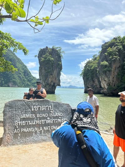 A man and woman pose for a photo in front of a stone monument that reads "JAMES BOND ISLAND" and Thai script. They are standing in shallow water. In the background, a tall, rocky island covered in greenery rises from the sea. The sky is blue with scattered clouds. Other people are visible in the background, including a man in a white shirt and sunglasses and another man in an orange shirt. The monument is located on a sandy beach. The location is James Bond Island in Phang Nga Bay, Thailand.