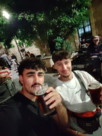 The image depicts two males in the foreground, holding glasses of dark beer, within an outdoor dining or bar establishment located in West Jerusalem, Israel. The scene is illuminated by artificial light, indicating evening or nighttime.

The male positioned on the left has dark, curly hair and a prominent mustache, wearing a dark short-sleeved shirt. He holds a glass of dark beer in his right hand, raised towards the camera, and looks directly at the lens with his mouth slightly ajar. The male on the right, partially visible, has brown wavy hair, wears a white short-sleeved t-shirt, a silver chain necklace, and a dark fanny pack around his waist. He smiles towards the camera while holding a glass of dark beer in his left hand.

The background reveals a social outdoor setting with additional patrons seated at dark tables and chairs. Green foliage from trees or bushes is visible overhead and to the sides. Stone-colored building walls, characteristic of Jerusalem architecture, are present, along with a large window opening. Artificial light sources, including a visible streetlight in the upper left, illuminate the environment. Other individuals in the mid-ground include one person wearing a white baseball cap and a dark jacket with crossed arms, and another in a red and white striped shirt.
